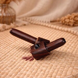May include: Two-piece, dark brown wooden pipe with a black stone accent. The pipe is angled and rests on a woven surface. Dried plants and a glass vase are in the background.
