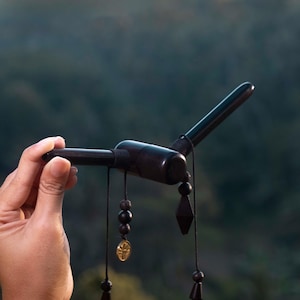 May include: A dark brown wooden pipe with a curved design, held in a hand. The pipe features dangling black beads, a gold-colored charm, and a geometric pendant. The background is a blurred view of a green forest.