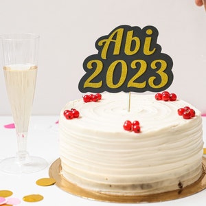 May include: A white cake with red berries and a black and gold glitter cake topper that says "Abi 2023".