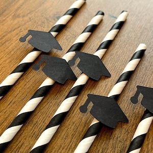 May include: Close-up of graduation-themed paper straws. The straws are black and white striped with small black graduation cap cutouts attached. The straws are arranged diagonally on a wooden surface, perfect for a graduation party.