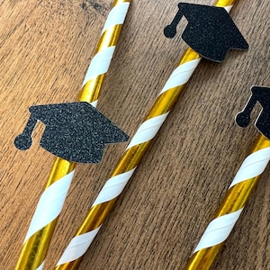 May include: Three gold and white striped drinking straws with black graduation cap cutouts attached. The straws are on a wood background.