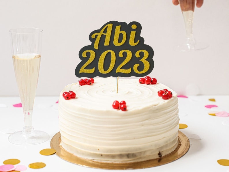 May include: A white cake with red berries and a black and gold glitter cake topper that says "Abi 2023".