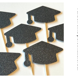 May include: Glittery black graduation cap cupcake toppers on wooden sticks. The toppers are arranged on a white surface. The text on the right side of the image says "Farben sind personalisierbar".