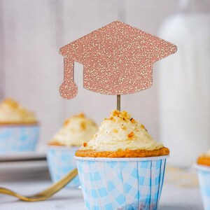 May include: Cupcakes with white frosting and a graduation cap topper. The cupcakes are in blue and white checkered paper cups. The graduation cap is rose gold glitter.