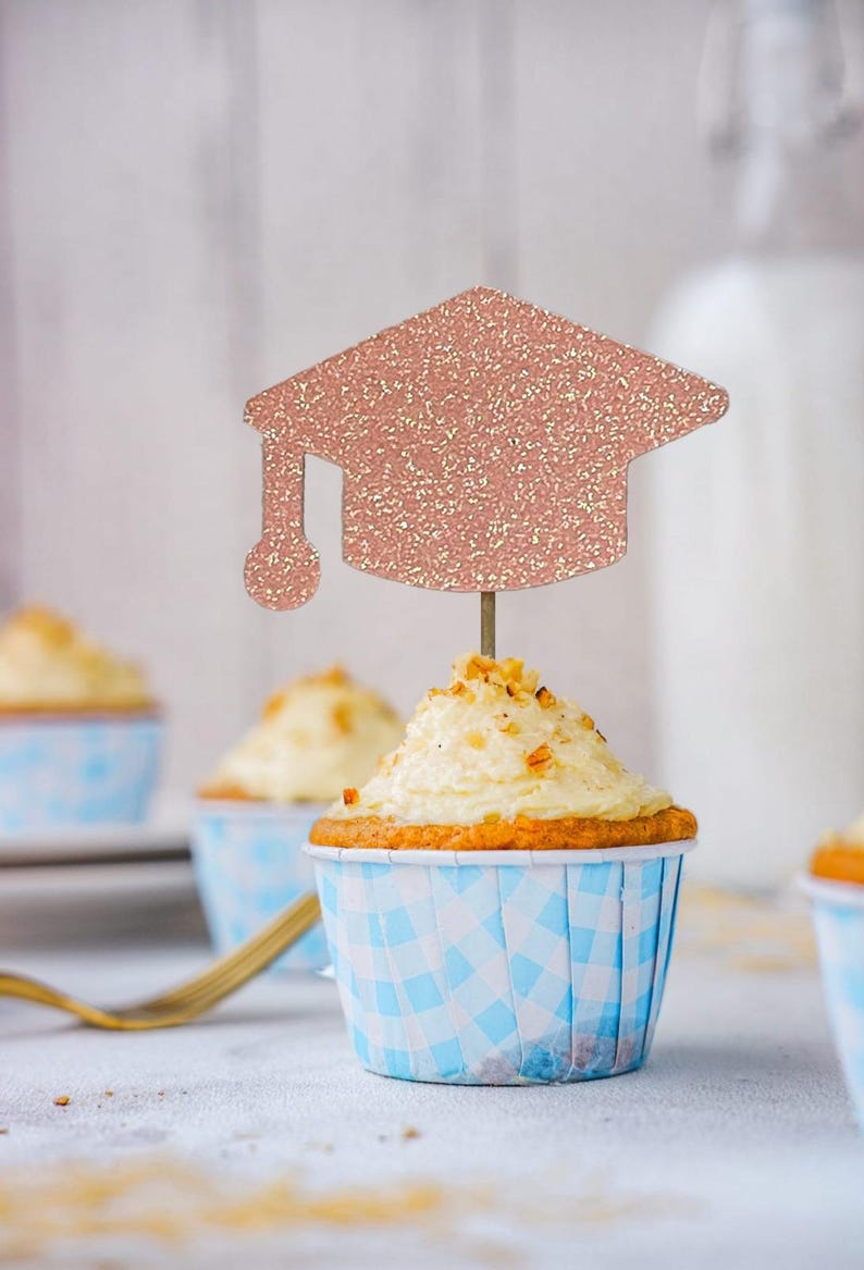 May include: Cupcakes with white frosting and a graduation cap topper. The cupcakes are in blue and white checkered paper cups. The graduation cap is rose gold glitter.