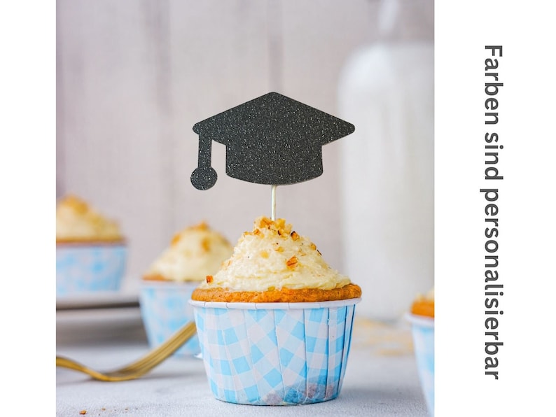 May include: A cupcake with white frosting and a black glitter graduation cap topper. The cupcake is in a blue and white checkered liner. The text on the right side of the image says "Farben sind personalisierbar".