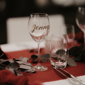May include: A clear glass wine glass with the name "Jenny" written on it in gold lettering. The glass is sitting on a red tablecloth with green leaves.