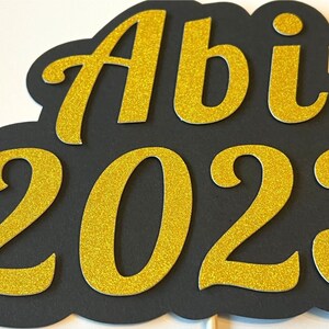 May include: Black and gold glitter graduation sign with the text "Abi 2023".