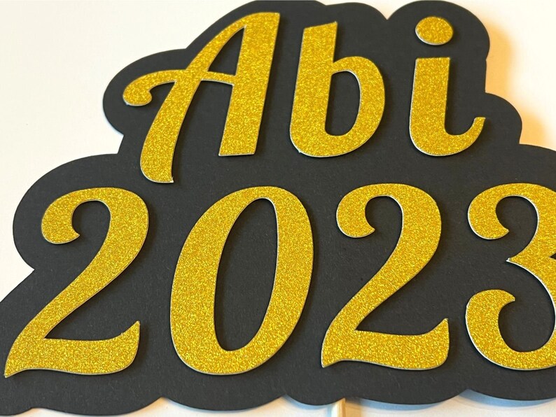 May include: Black and gold glitter graduation sign with the text "Abi 2023".