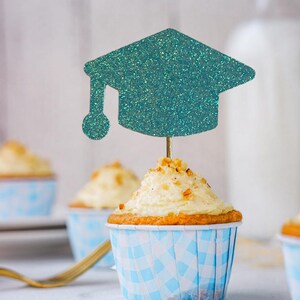 May include: A cupcake with white frosting and a teal glitter graduation cap topper. The cupcake is in a blue and white checkered paper cup. Other cupcakes are in the background.
