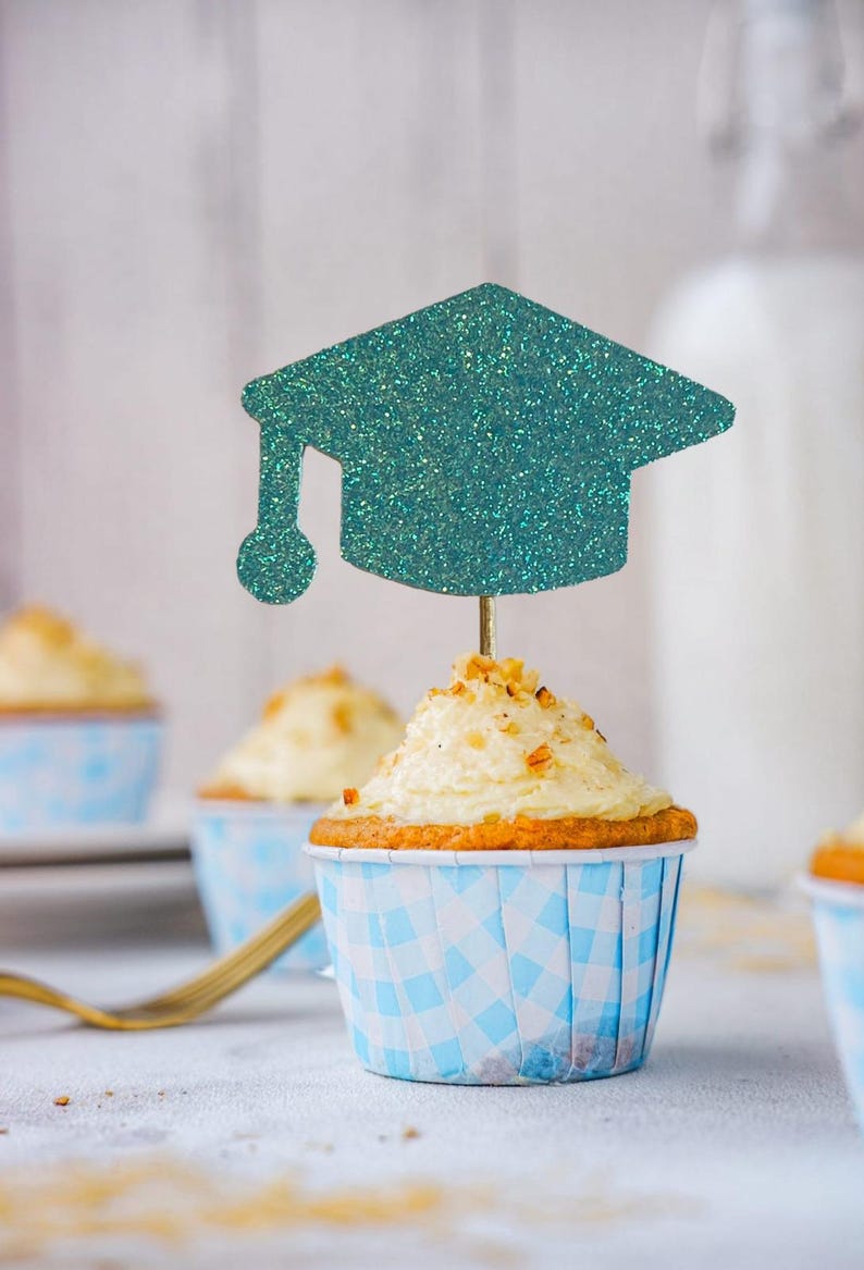 May include: A cupcake with white frosting and a teal glitter graduation cap topper. The cupcake is in a blue and white checkered paper cup. Other cupcakes are in the background.