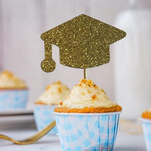 May include: A cupcake with white frosting and a gold glitter graduation cap topper. The cupcake is in a blue and white checkered liner. Other cupcakes are in the background.