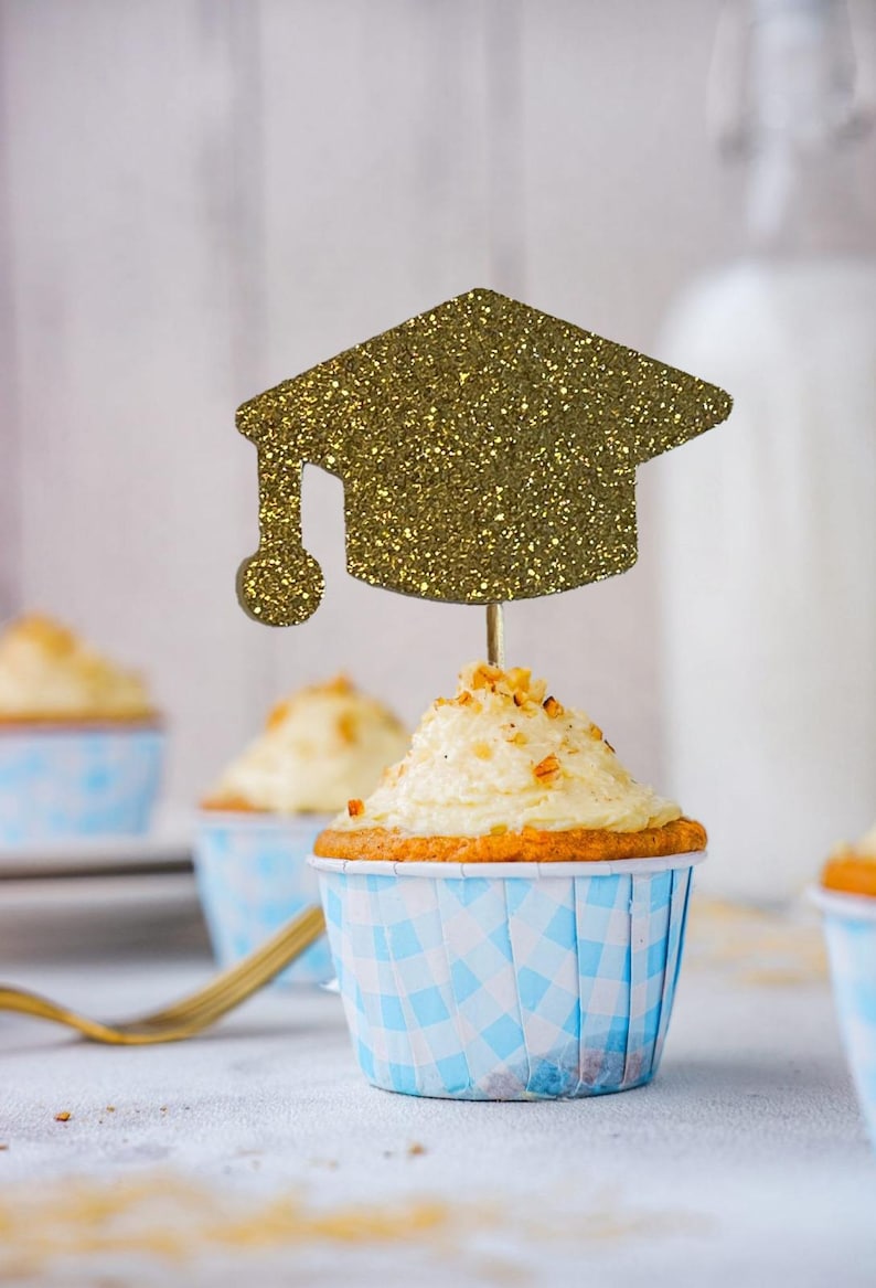 May include: A cupcake with white frosting and a gold glitter graduation cap topper. The cupcake is in a blue and white checkered liner. Other cupcakes are in the background.