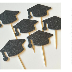 May include: Black glitter graduation cap cupcake toppers on wooden sticks. The image shows several toppers arranged on a white surface. The text on the right side of the image says "Colors are customizable".