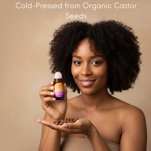 May include: A woman holds a small roller bottle of oil and a handful of castor seeds. The bottle has a colorful label with a gradient design. The text "Cold-Pressed from Organic Castor Seeds" is at the top of the image.
