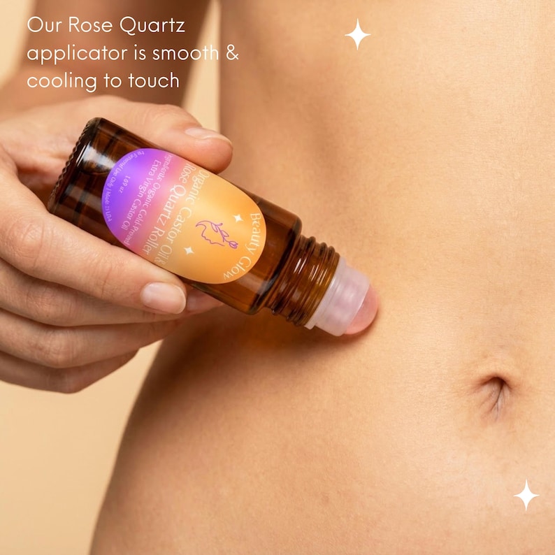 May include: A brown glass roller bottle with a pink rose quartz applicator is being used on skin. The bottle has a label with text that reads "Organic Castor Oil Rose Quartz Roller". The text "Our Rose Quartz applicator is smooth & cooling to touch" is also visible.