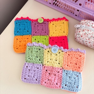 May include: Two rectangular crochet pouches made from colorful granny squares. The top pouch features blue, orange, purple, lime green, yellow, and hot pink squares with a lime green button. The bottom pouch has light green, yellow, peach, lavender, pink, and light blue squares with a light blue button.