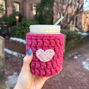May include: A bright magenta crochet cup cosy featuring a light pink heart appliqué centred on the front. The cosy is wrapped around a clear glass jar containing a light-coloured beverage with a silver metal straw protruding from the top.