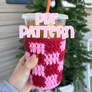 May include: A clear plastic iced coffee cup with a pink and red crochet cozy. The cozy has a checkered pattern. The cup has a pink and orange straw. The text "PDF PATTERN" is overlaid on the image.