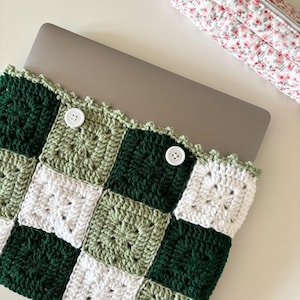 Puede incluir: Una funda para portátil de punto de crochet en verde, blanco y verde salvia con botones blancos. La funda tiene un patrón de cuadros.