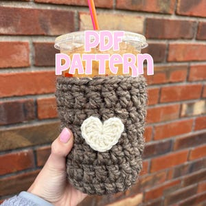 May include: A brown crochet coffee cup cozy with a white heart applique. The cozy is wrapped around a clear plastic cup with a pink and orange straw. The words "PDF PATTERN" are in pink letters across the cup.
