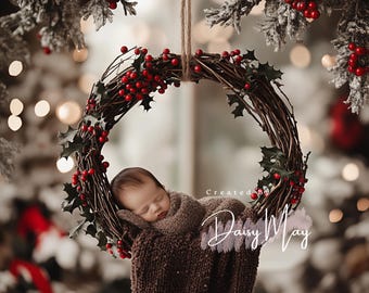 May include: A newborn baby sleeping in a brown knitted wrap inside a rustic twig wreath decorated with red berries and green holly leaves. The wreath is hanging from a white string. The background is blurred and features a Christmas tree with lights.