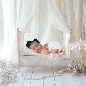 May include: A newborn baby sleeping in a white crib with a white canopy. The crib is decorated with white lace and pearls. The baby is wearing a white dress and a white hat. The background is a white wall with white curtains.