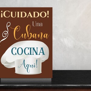 Puede incluir: Un letrero marrón con texto blanco que dice "¡CUIDADO! Una Cubana COCINA Aqui!"  El letrero también presenta un gorro de chef blanco con un borde azul verdoso.