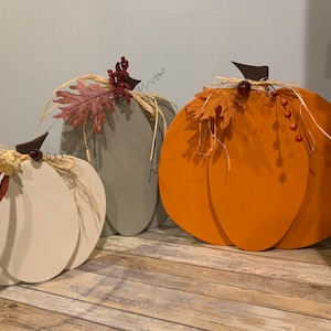 May include: Three wooden pumpkins in varying sizes and colors, perfect for fall decor. The pumpkins are painted in white, gray, and orange. Each pumpkin is adorned with decorative stems, raffia, and fall-themed embellishments. They are displayed on a rustic wooden surface.