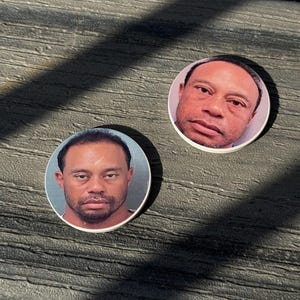 May include: Two round coasters featuring a portrait of a man with dark hair and a beard. The image is a close-up of the man's face, with a neutral expression. The coasters are white with a glossy finish, and are placed on a textured, gray surface.
