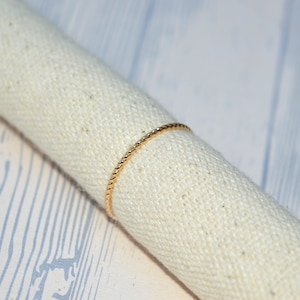 Könnte beinhalten: Ein dünner goldener Ring mit einem gedrehten Design. Der Ring liegt auf einer weißen Stoffoberfläche.