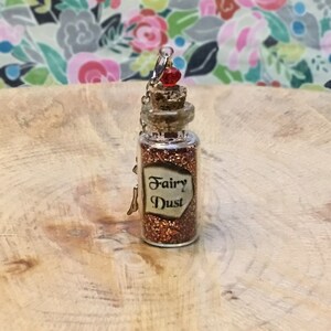May include: A small glass bottle with a cork stopper and a silver chain. The bottle is filled with glittery gold and red fairy dust. The label on the bottle reads "Fairy Dust".