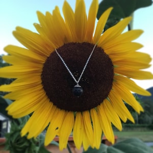 May include: A silver necklace with a dark, round pendant is displayed against a vibrant yellow sunflower. The pendant has a textured surface and is suspended from a delicate chain. The sunflower's center is a deep brown.