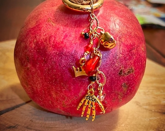 May include: A ripe red pomegranate with a gold-colored charm attached. The charm features a gold ring, a crown, a heart, a hand, and red and black beads. The pomegranate is on a wooden surface.