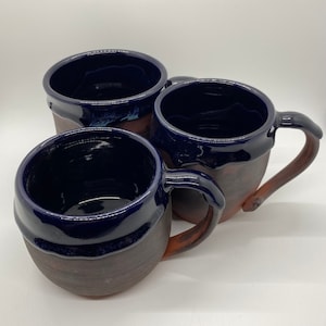 Könnte beinhalten: Drei handgefertigte Keramikbecher mit dunkelblauer Glasur und braunem Tonkörper. Die Becher haben eine abgerundete Form und einen breiten Rand.