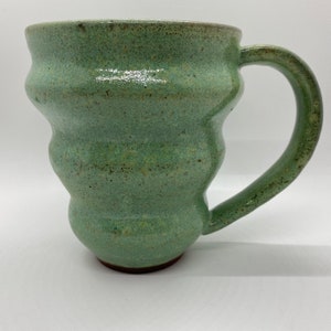 May include: A green ceramic mug with a textured, speckled glaze and a rounded, flared body. The mug has a large handle and a slightly uneven rim.