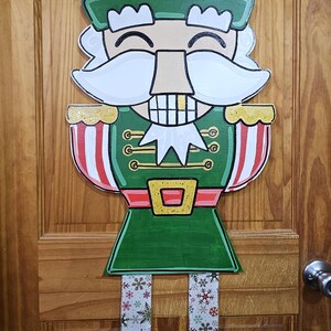 May include: A wooden Christmas nutcracker door hanger. The festive decoration features a smiling nutcracker in a green uniform with red and white striped accents. The legs have a snowflake pattern.