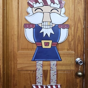 May include: A patriotic wooden door decoration in the shape of a nutcracker. The nutcracker is dressed in a blue uniform with a red and white striped hat and boots. The hat is decorated with white stars.
