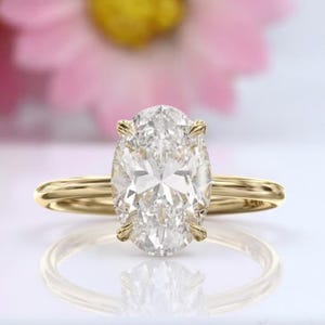 Puede incluir: Un anillo de compromiso dorado con un gran diamante de talla ovalada. El anillo está sobre una superficie blanca, con flores rosas en el fondo. El diamante es claro y brillante.