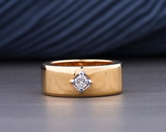 May include: A wide, polished yellow gold band ring featuring a single princess-cut diamond set in a square, silver-toned metal bezel at the center. The ring has a smooth, flat surface and a consistent width throughout its circular design.