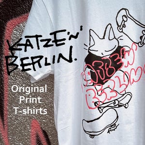 May include: A white t-shirt featuring a black line drawing of a cat on a skateboard, with additional skateboards and pink graffiti-style text. The shirt includes the printed text KATZEN BERLIN and Original Print T-shirts in black lettering.