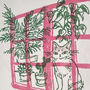 May include: A white art print featuring a green ink illustration of a cat sitting next to several potted plants in front of a pink window frame. The artwork includes a signature in gold ink at the bottom left corner, dated 2026, on a white background.