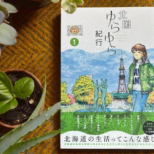 May include: A book cover with Japanese text and illustrations. The cover features a young woman in a green jacket and blue jeans, standing in front of a cityscape with a tower. The title is in Japanese, with the number 1 in a green circle.