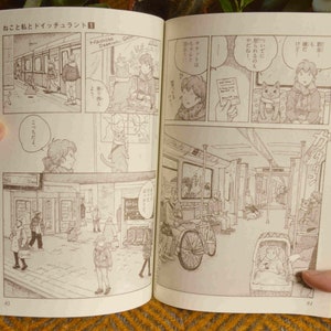 Puede incluir: Un manga con una portada marr&oacute;n e ilustraciones en blanco y negro. El manga est&aacute; abierto en una p&aacute;gina con una escena de personas en un tren. El texto en la p&aacute;gina est&aacute; en japon&eacute;s.