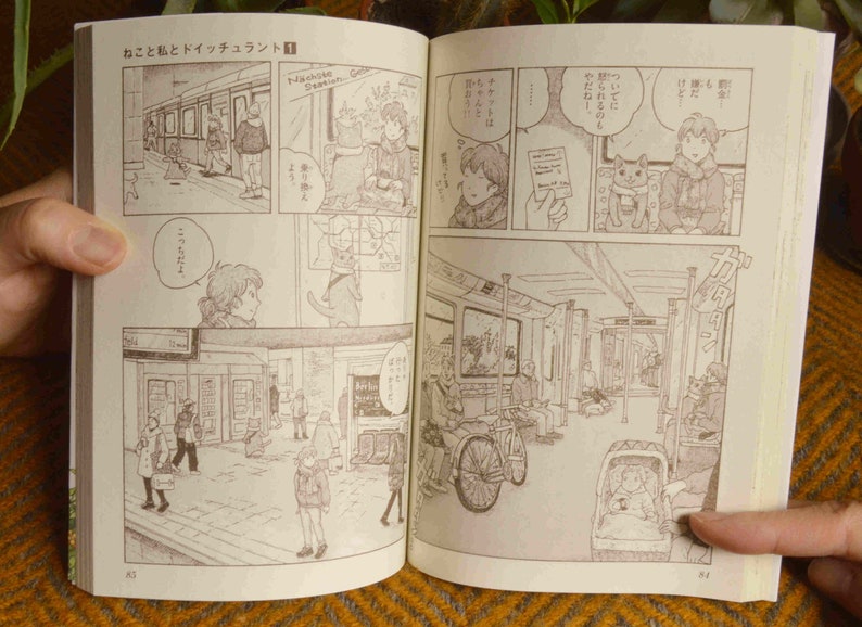 Puede incluir: Un manga con una portada marr&oacute;n e ilustraciones en blanco y negro. El manga est&aacute; abierto en una p&aacute;gina con una escena de personas en un tren. El texto en la p&aacute;gina est&aacute; en japon&eacute;s.