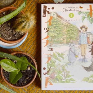 Puede incluir: Un libro de manga japon&eacute;s titulado "Meine Katze und ich in Deutschland" con un gato azul y una persona caminando por una calle. La portada del libro est&aacute; ilustrada en estilo acuarela con una gama de colores azul claro, verde y marr&oacute;n.