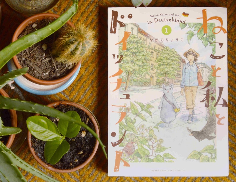 Puede incluir: Un libro de manga japon&eacute;s titulado "Meine Katze und ich in Deutschland" con un gato azul y una persona caminando por una calle. La portada del libro est&aacute; ilustrada en estilo acuarela con una gama de colores azul claro, verde y marr&oacute;n.