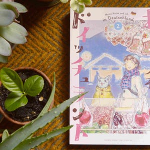Puede incluir: Un libro de manga japon&eacute;s titulado "Meine Katze und ich in Deutschland" con el n&uacute;mero 2 en la portada. La portada presenta a una ni&ntilde;a con cabello casta&ntilde;o que lleva una bufanda azul y un sombrero blanco, un gato que lleva una chaqueta amarilla, y un fondo de un paisaje nevado con un puente.