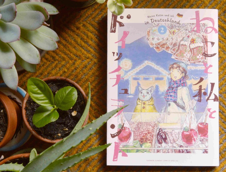 Puede incluir: Un libro de manga japon&eacute;s titulado "Meine Katze und ich in Deutschland" con el n&uacute;mero 2 en la portada. La portada presenta a una ni&ntilde;a con cabello casta&ntilde;o que lleva una bufanda azul y un sombrero blanco, un gato que lleva una chaqueta amarilla, y un fondo de un paisaje nevado con un puente.