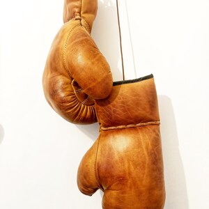May include: A pair of brown leather boxing gloves hanging from a string. The gloves are worn and have a distressed look.